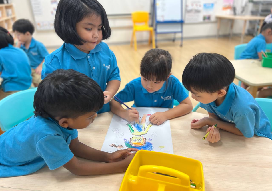 Four children in blue uniforms color a drawing together at a classroom table, with crayons nearby.